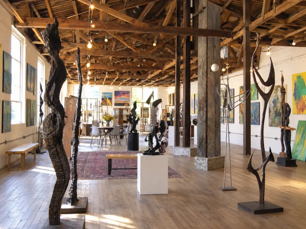 A light-filled gallery with metal sculptures and paintings under exposed timber beams